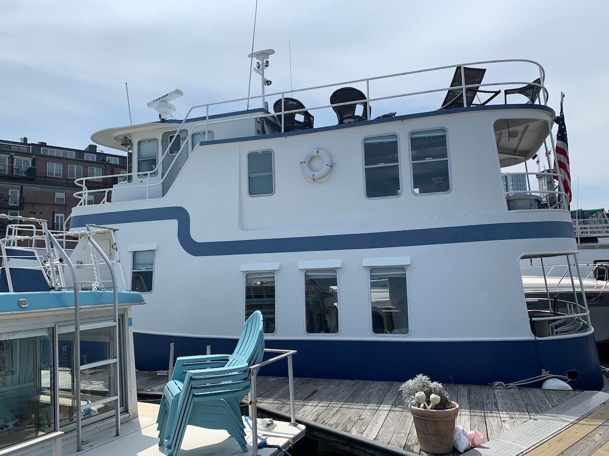Un grand bateau blanc et bleu était amarré dans une marina. Il était doté d’une terrasse couverte, d’une bouée de sauvetage et d’une petite plante en pot.