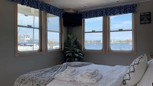 A bedroom with a bed, two windows with a view of the water, a plant, and a sign on the wall.