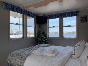 A bedroom with a bed, two windows with a view of the water, a plant, and a sign on the wall.