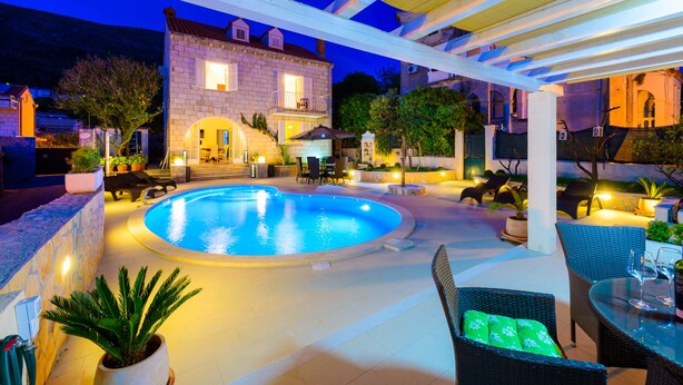 A pool area with a stone house in the background, outdoor furniture, and a pergola.