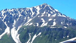 A mountain with snow patches and a clear sky. There are two white buildings with a sloped roof, one with a wooden facade. - Howling Wolf Resort!  (Seward)