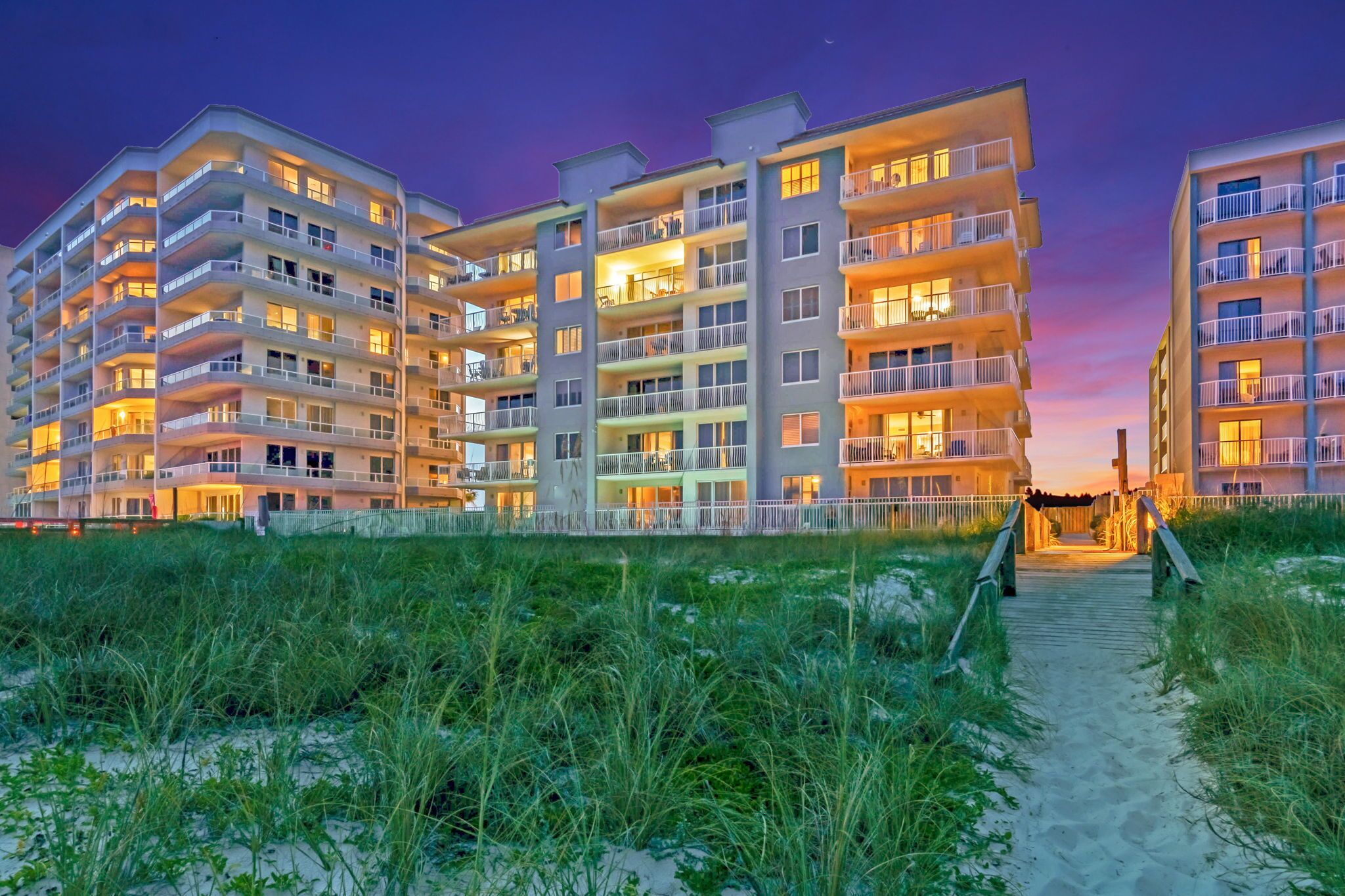 A multi-story hotel with balconies, a sandy pathway leading to the entrance, and a beach in the foreground.