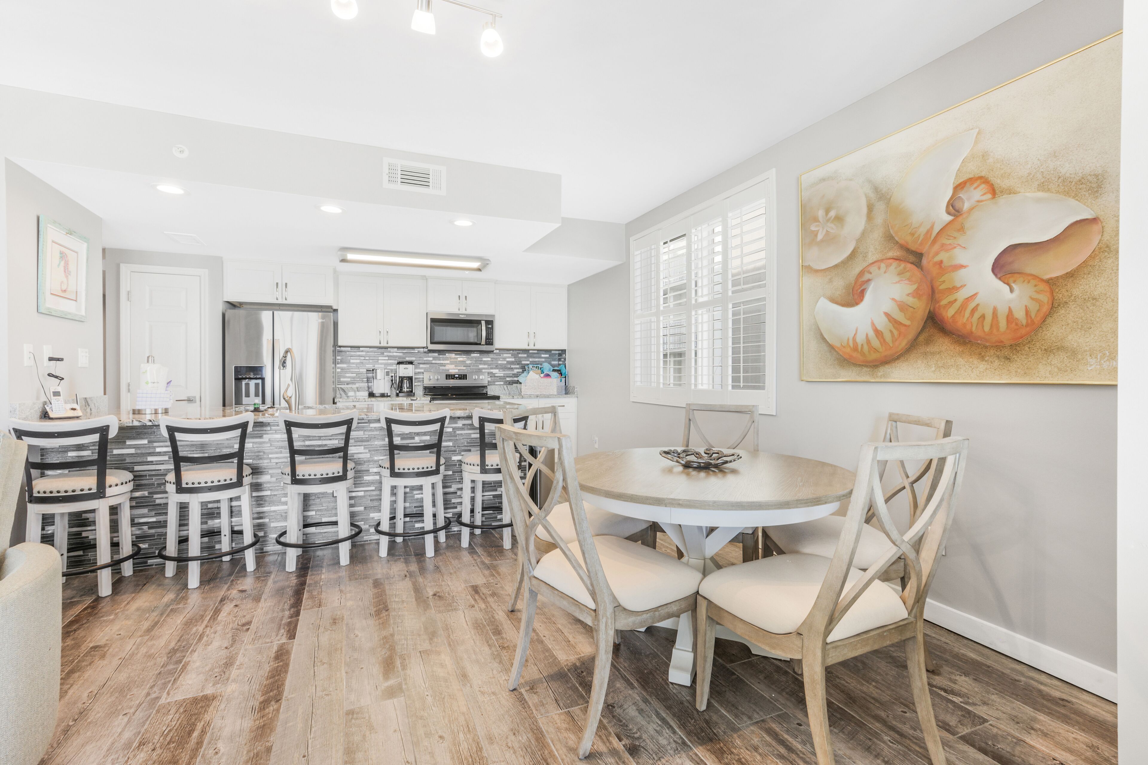 A modern kitchen with white cabinets, a stainless steel refrigerator, and a microwave. There is a dining area with a round table and chairs.