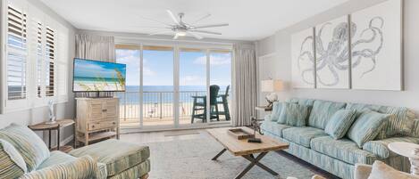 A spacious living room with a large TV, a ceiling fan, and a view of the ocean through large windows.
