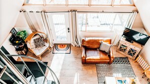 A cozy loft-style living space with a high ceiling, a large window, a leather sofa, a TV, and a staircase.