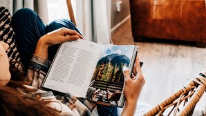 A person reading a book in a wicker chair in a room with a bed and a TV in the background.