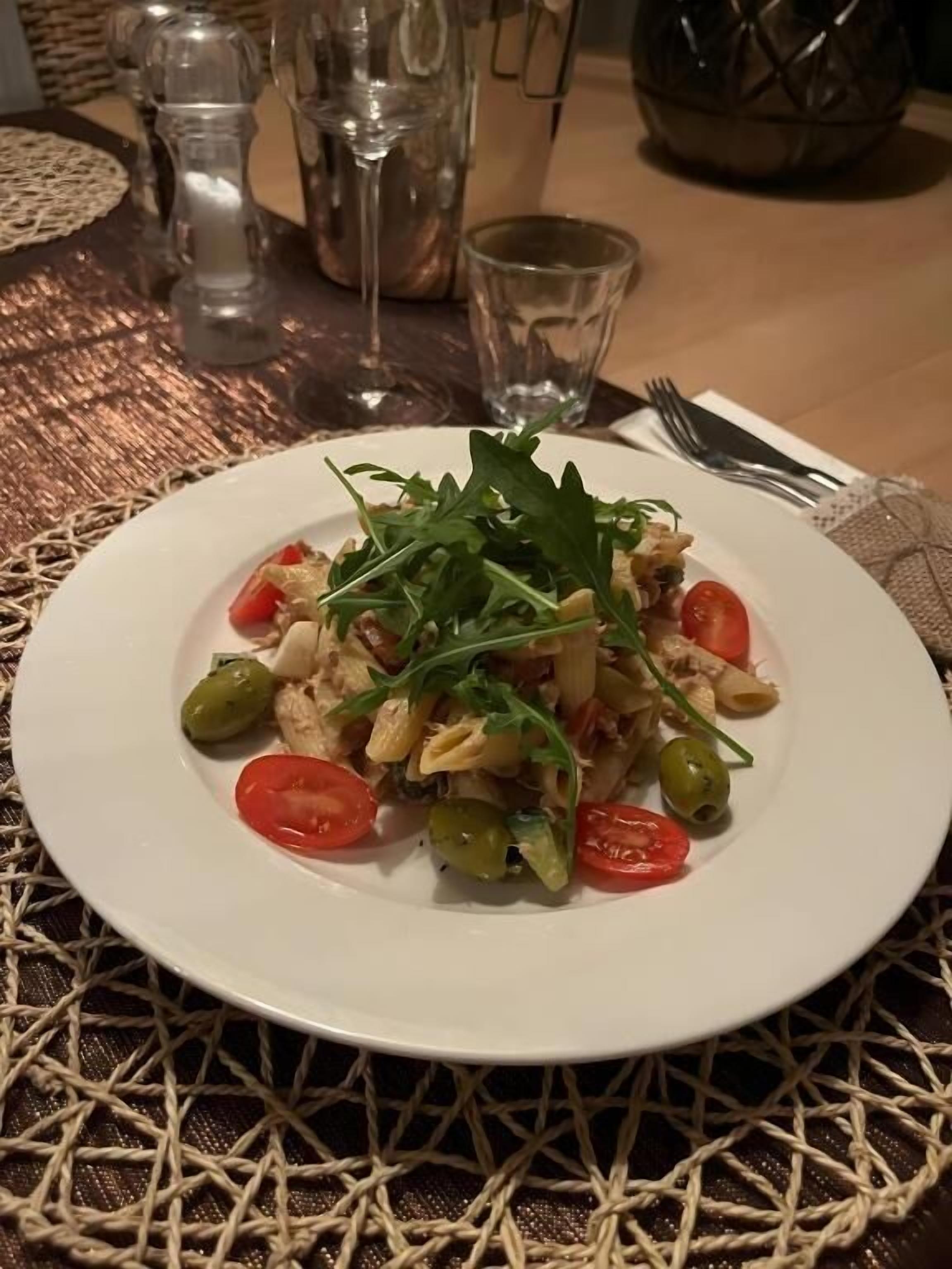 En tallrik pasta med körsbärstomater, gröna oliver och rucola, serverad på en vävd bordsduk.