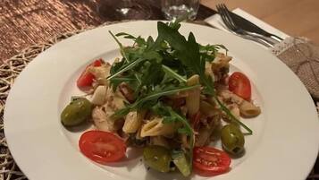 En tallrik pasta med körsbärstomater, gröna oliver och rucola, serverad på en vävd bordsduk.