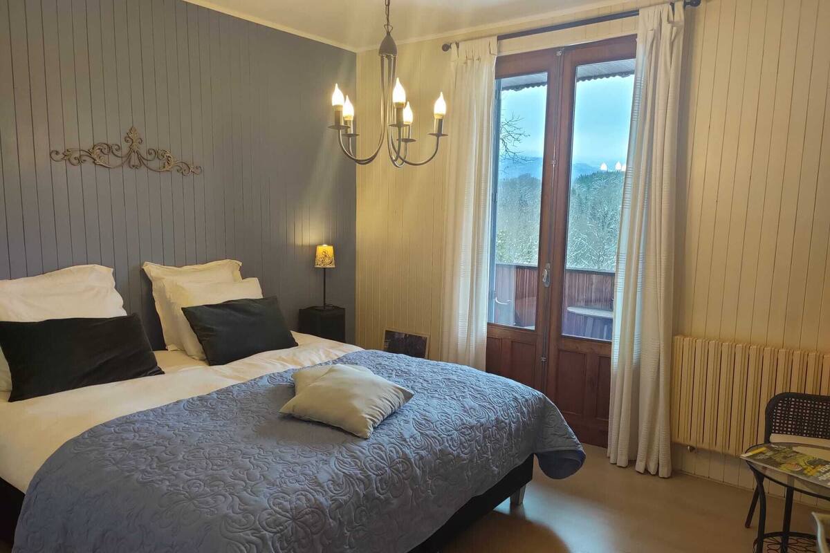A bedroom with a bed, a chandelier, a nightstand, a window with curtains, and a view of the outdoors.
