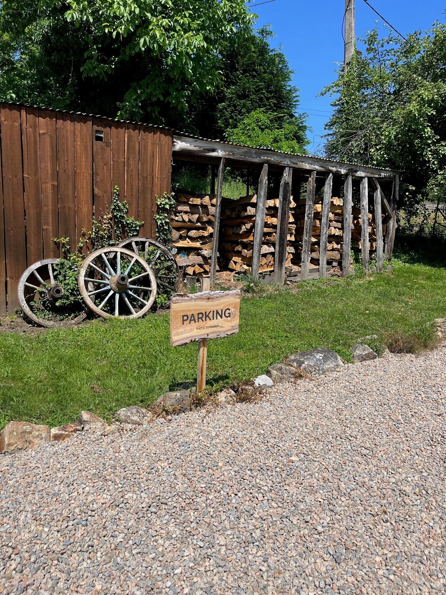 Un panneau en bois sur lequel est écrit « PARKING », placé devant une structure rustique en bois, avec une pile de bois de chauffage et deux vieilles roues de charrette.