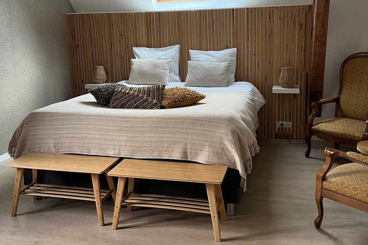 A bedroom with a sloped ceiling, a skylight, a bed with pillows, a bedside table, a chair, and a wooden bench.
