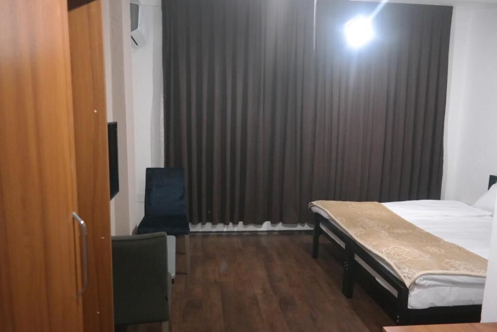 A hotel room with a wooden floor, a bed with a beige bedspread, a desk with a chair, and a large window with curtains.