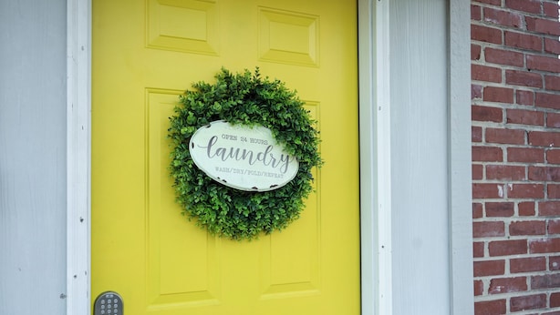 A yellow door with a green wreath and a sign that reads "Open 24 Hours Laundry Wash/ Dry/Fold/ Repeat". - Mid Century Remodeled Suite in Midtown – 20A (Mobile)