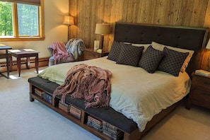 A bedroom with a large bed, bedside lamps, a wooden headboard, a bench with books, and a window with a view of trees.