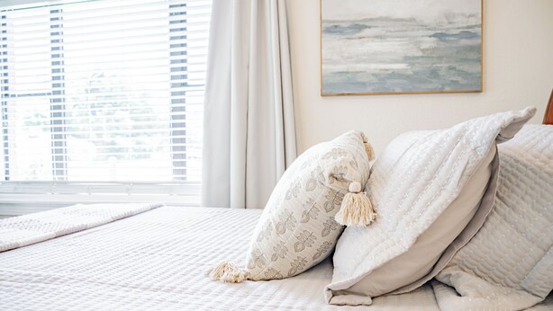Ein ordentlich bezogenes Bett mit gemusterten Kissen und einer gesteppten Decke, ein Fenster mit Jalousien und ein abstraktes Bild an der Wand.