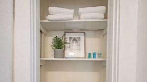 Ein weißer Kleiderschrank mit Regalen, auf denen gefaltete Handtücher, ein Blumentopf, Toilettenartikel und ein Korbwäscheschrank stehen.