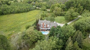 Une grande maison avec une piscine, entourée d’une forêt dense et de champs.