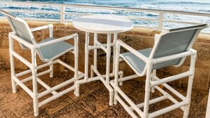 Un patio con dos sillas de mimbre blancas y una pequeña mesa redonda, con vista a una playa con olas y una costa lejana.