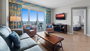 Ein Wohnzimmer mit einer Couch, Sesseln, einem Couchtisch und einem Fernseher. Es gibt einen Balkon mit Blick auf den Ozean.