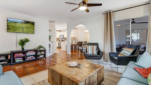 A spacious living room with a wooden coffee table, a TV mounted on the wall, and a ceiling fan. - Heated Pool + Game Room | Near Vanderbilt Beach (Naples)