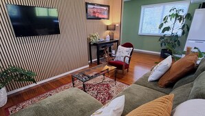 A modern living room with a flat-screen TV mounted on the wall, a dining table with chairs, and a large sofa with cushions. - Atherton Apartment with Views and Breakfast Hamper (Atherton)