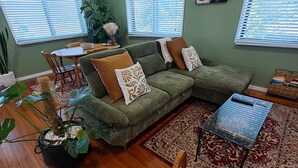 A living room with a green sectional sofa, a dining table with chairs, a coffee table, and a large window with blinds. - Atherton Apartment with Views and Breakfast Hamper (Atherton)