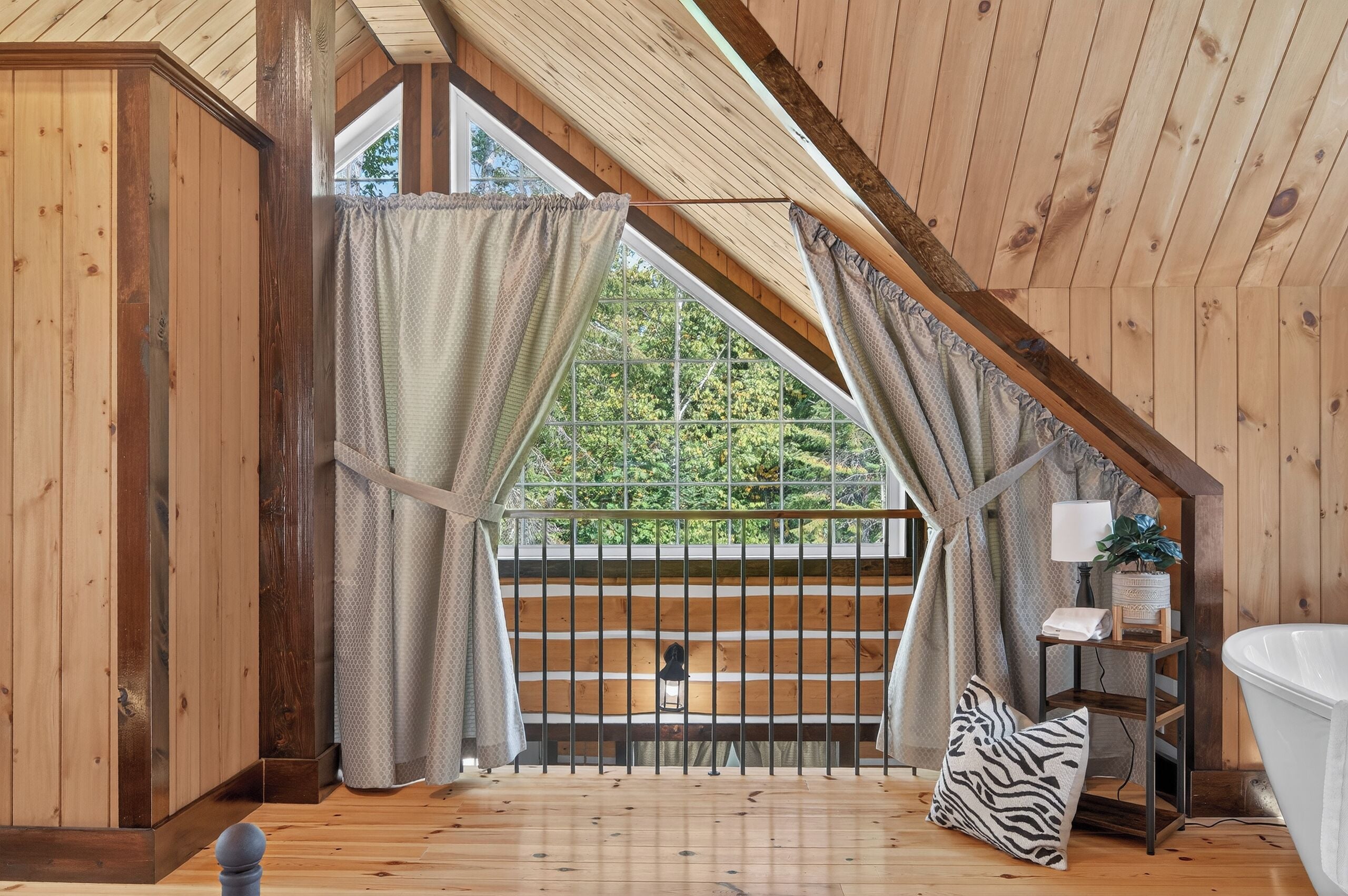 A wooden room with a sloped ceiling, a window with curtains, a small table with a lamp, a white bathtub, and a zebra-patterned pillow.