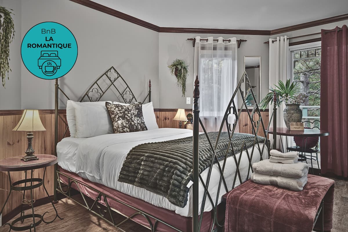 A bedroom with a four-poster bed, a bedside table with a lamp, a window with curtains, and a view of the outdoors.