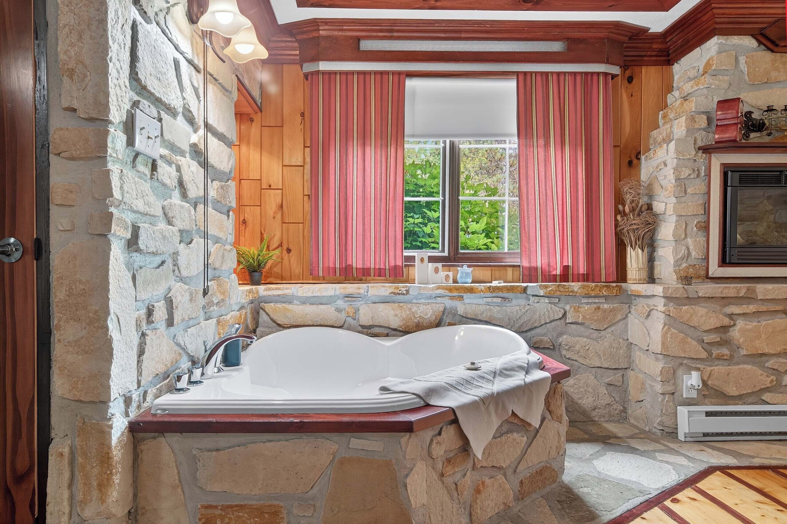 A stone-walled bathroom with a large bathtub, a window with curtains, and a fireplace.