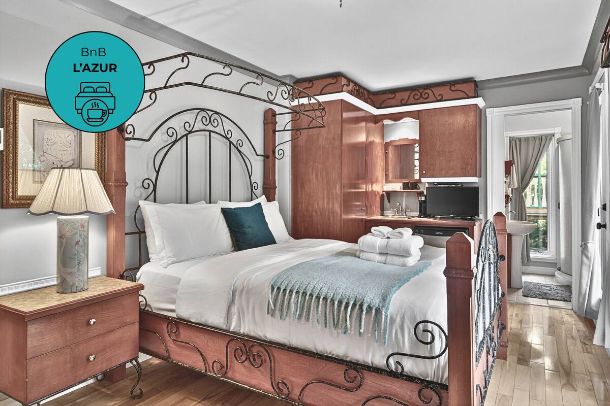 A bedroom with a four-poster bed, a bedside table with a lamp, a wooden wardrobe, a television, and a bathroom visible through an open door.