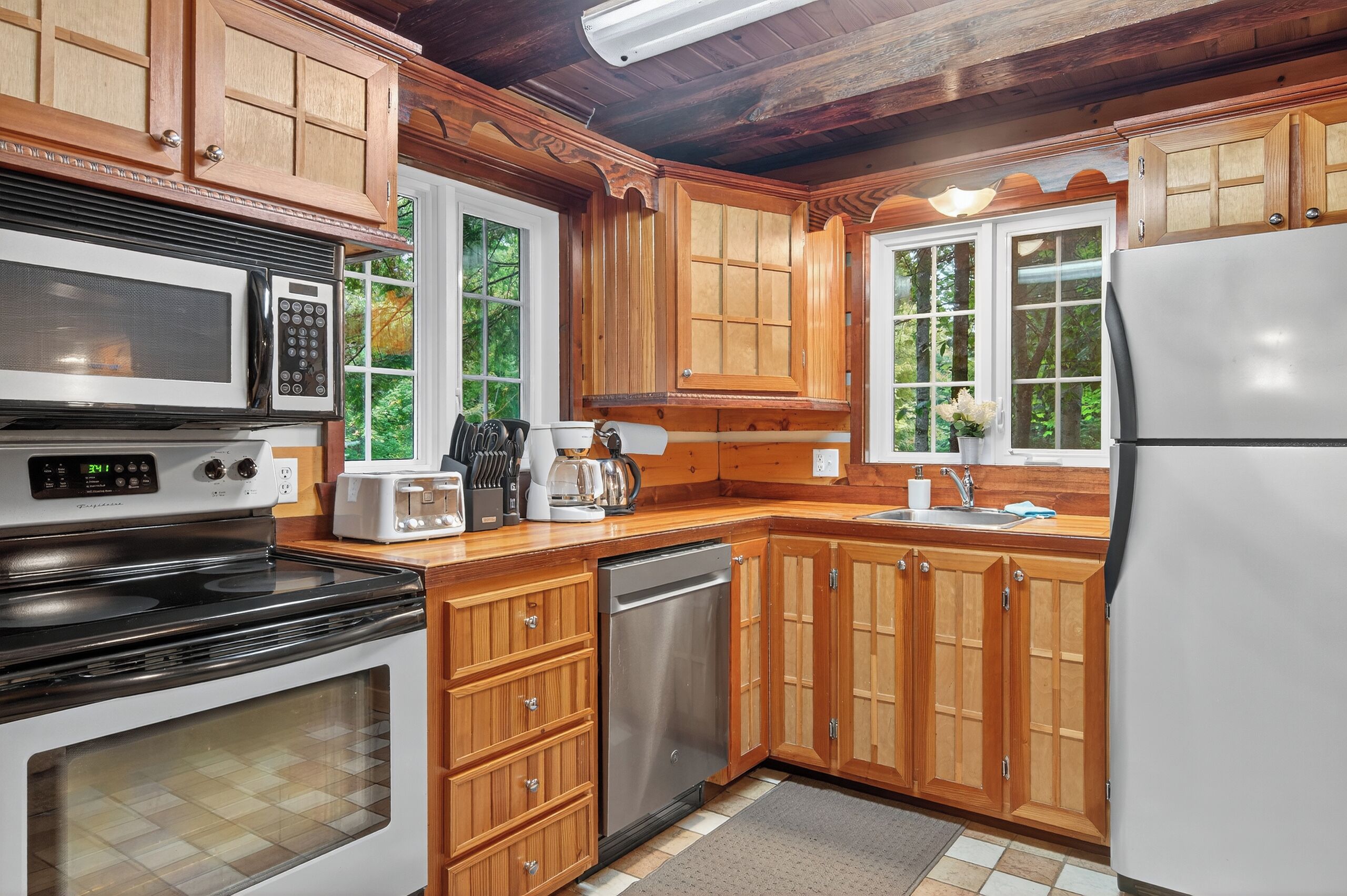 Une cuisine avec des meubles en bois, un réfrigérateur en acier inoxydable, un lave-vaisselle, un four à micro-ondes, une cuisinière et une fenêtre donnant sur des arbres.