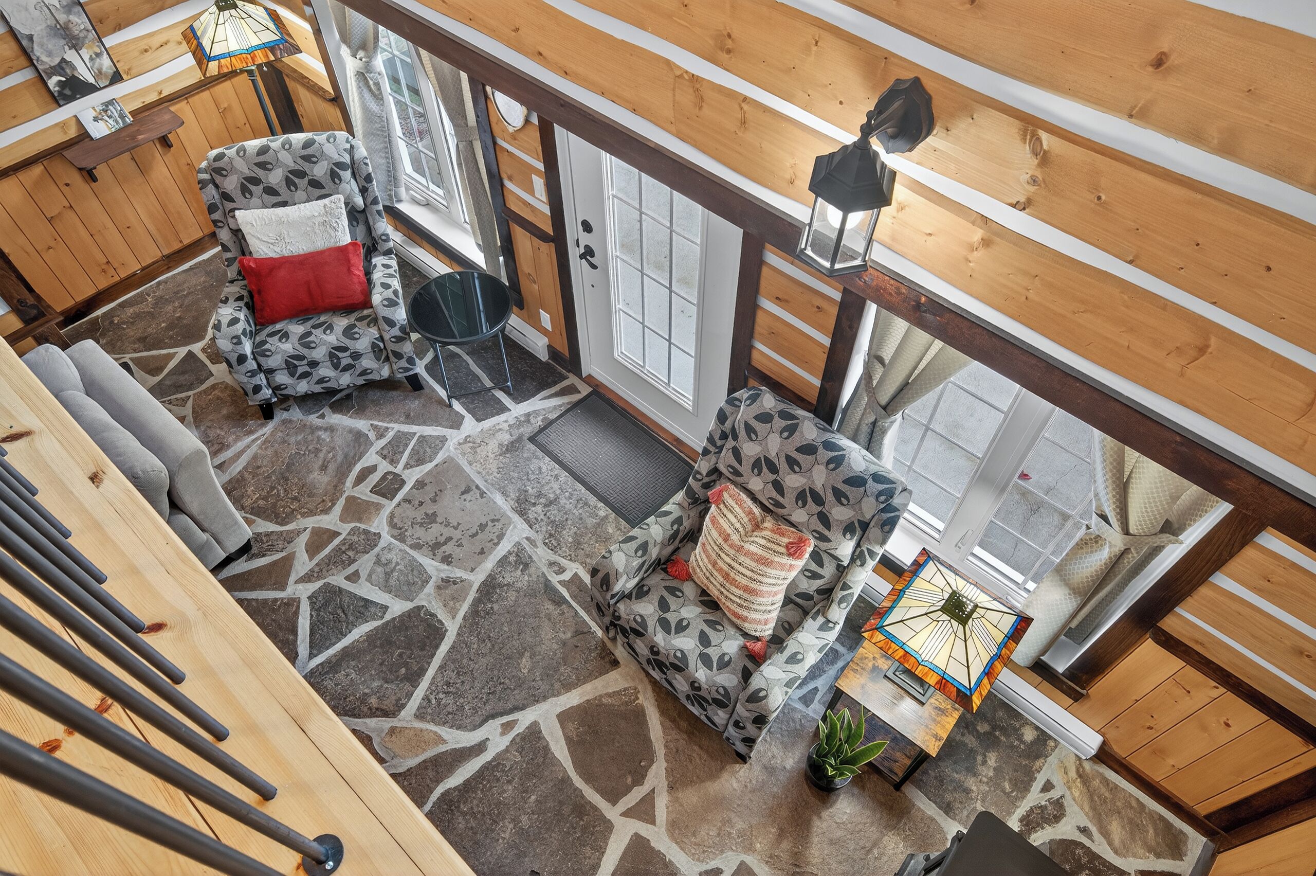 A spacious room with a stone-patterned floor, wooden walls, and a large window. There are two patterned armchairs with red and white cushions, a small table with a lamp, and a potted plant.