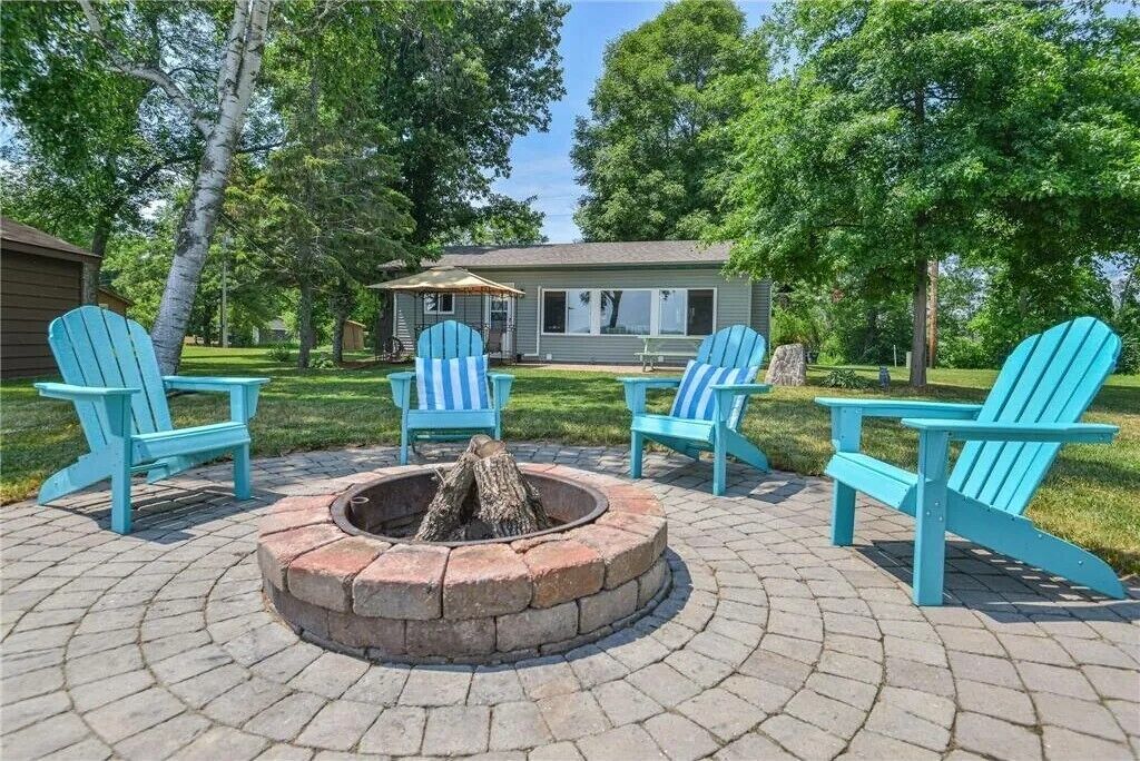 A patio area with a fire pit surrounded by four Adirondack chairs, a small building in the background, and a grassy lawn.
