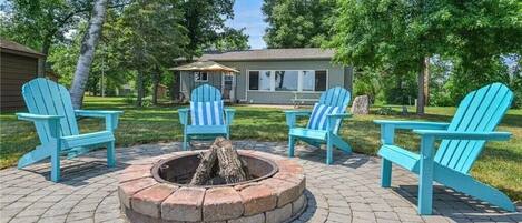 A patio area with a fire pit surrounded by four Adirondack chairs, a small building in the background, and a grassy lawn.