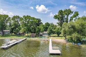 Una propiedad frente al lago con un muelle que se adentra en el agua, rodeada de árboles y casas.