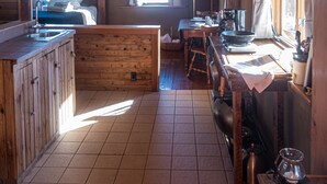Interior de una cabaña con techo de madera, una zona de cocina con estufa, una mesa de comedor y una cama con vista al exterior.