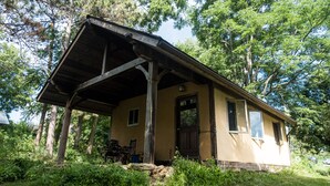 Una cabaña rústica con un porche de madera, una puerta y ventanas, rodeada de árboles y follaje.