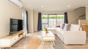 A modern living room with a flat-screen TV, a white sofa, a coffee table, and a large window with curtains. - Natural touch (Hamilton)