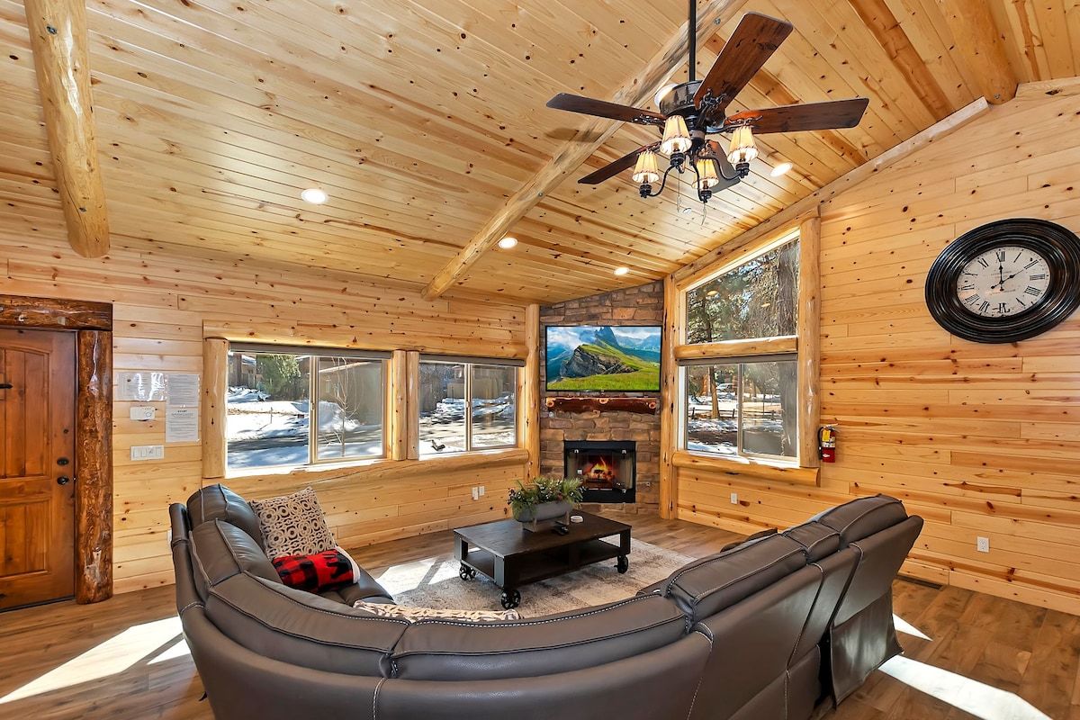 A cozy cabin interior with a fireplace, leather sofa, and large windows offering a view of the outdoors.