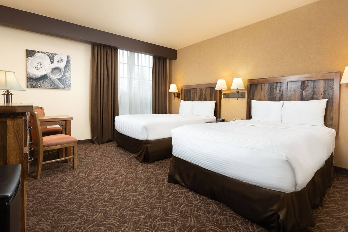 A hotel room with two beds, a desk, a chair, and a large window with curtains.