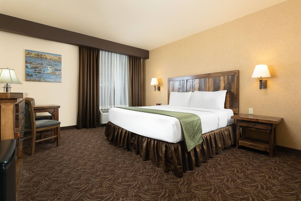 A hotel room with a large bed, two bedside tables with lamps, a desk with a chair, a large window with curtains, and a painting on the wall.