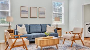 A living room with a blue sofa, wooden coffee table, and two chairs. There are framed pictures on the wall, a lamp, and a washing machine in the background.