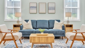 Ein Wohnzimmer mit einem blauen Sofa, einem Holztisch und zwei Sesseln. An der Wand hängen drei gerahmte Kunstwerke, und auf jedem Beistelltisch steht eine Lampe.