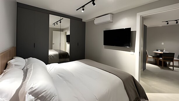 A modern hotel room with a large bed, a flat-screen TV mounted on the wall, and a separate dining area visible through an open door. - Noroeste - moderno e central (Brasília)