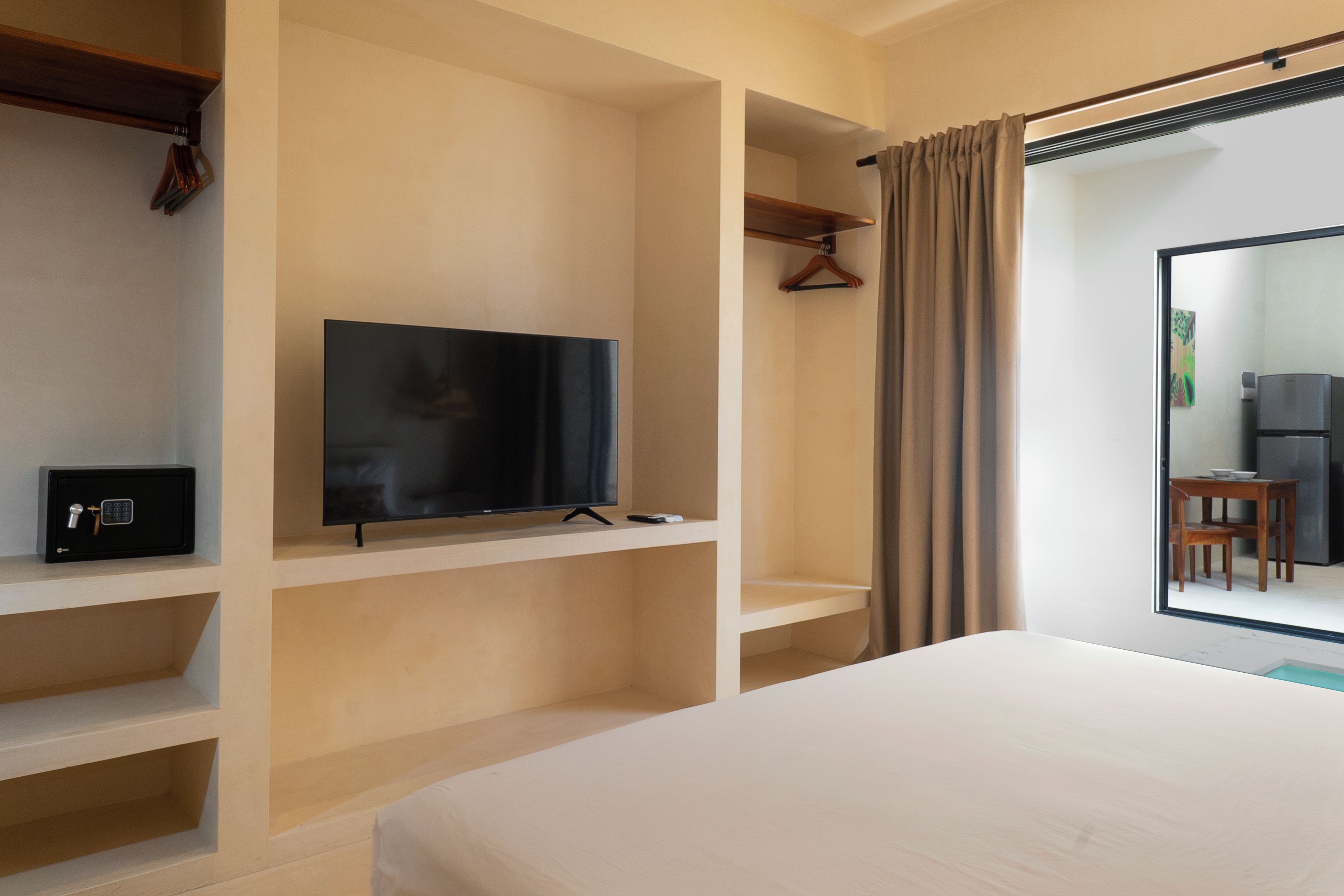 Habitación de hotel con cama, televisor en un estante, perchero y vista a un área de cocina.