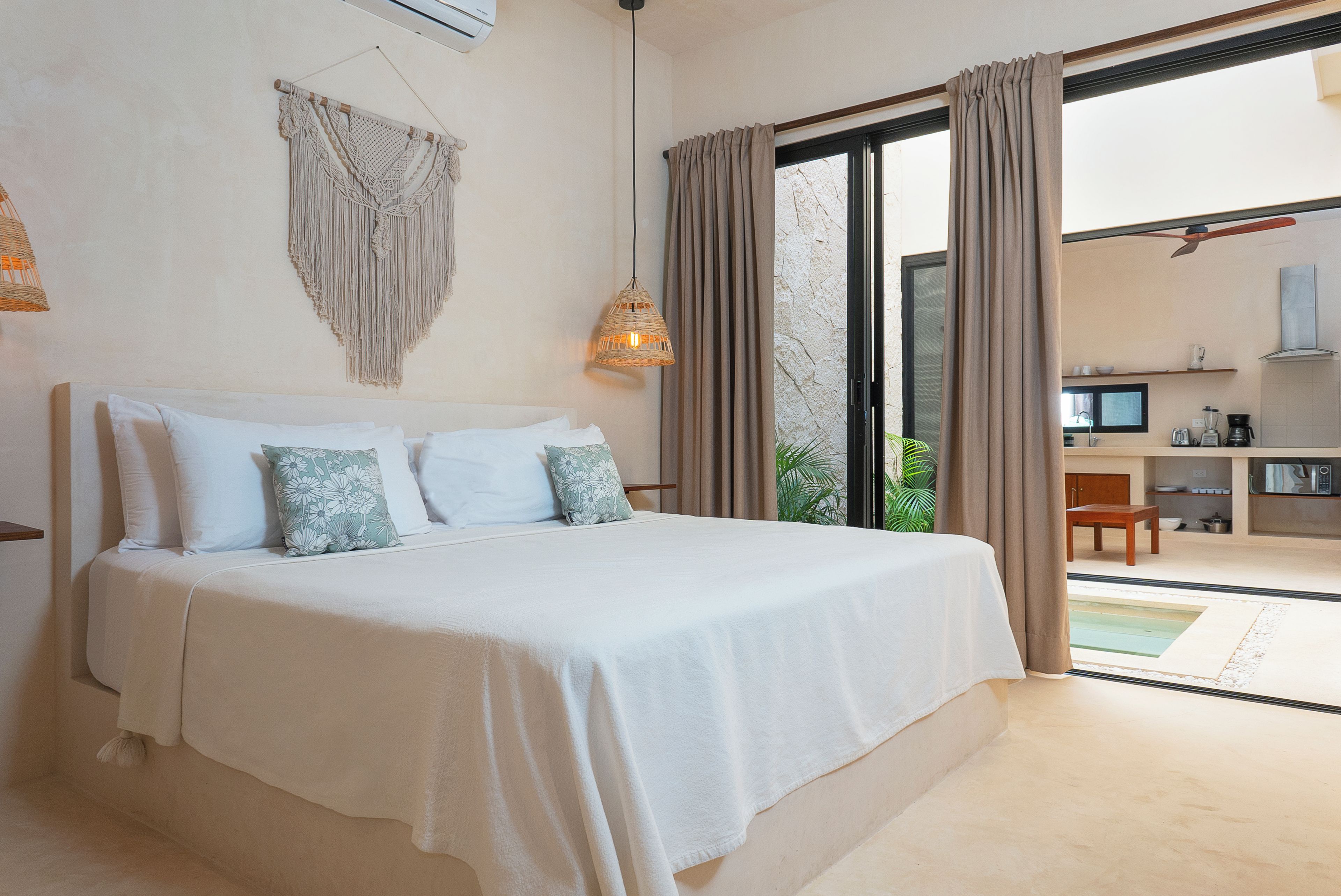 A bedroom with a large bed, a macrame wall hanging, and a view of a kitchen and outdoor area through a sliding door.