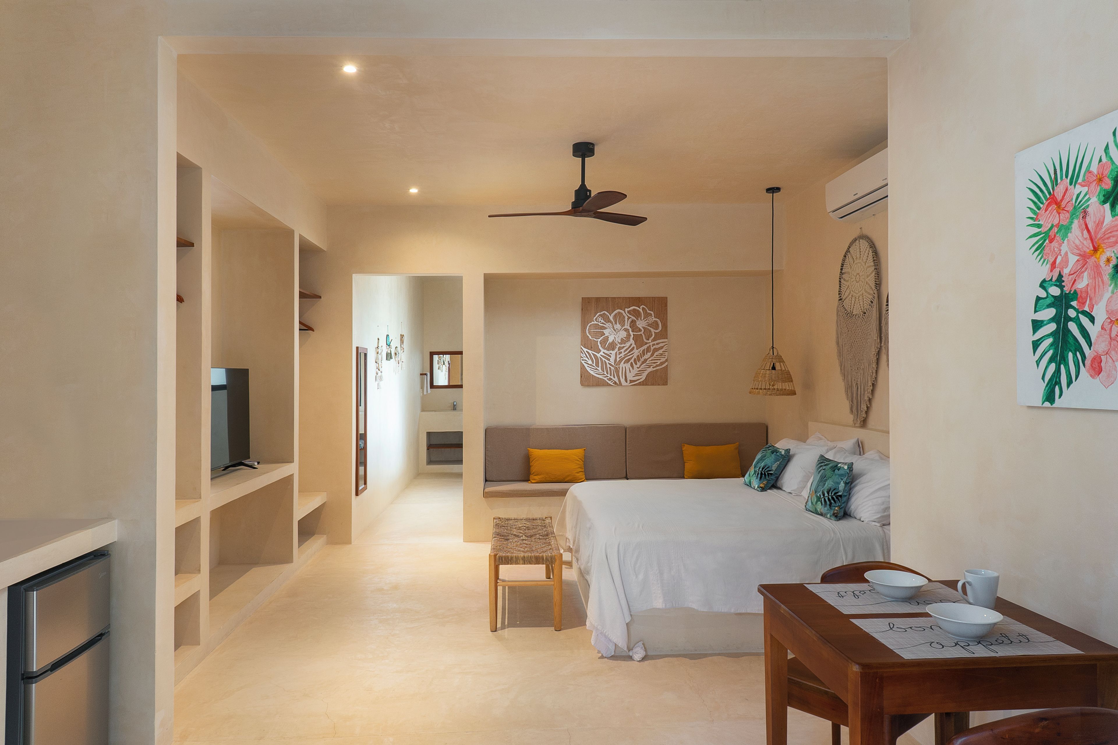 A hotel room with a bed, a desk with a bowl and mug, a ceiling fan, and a wall with a floral artwork.