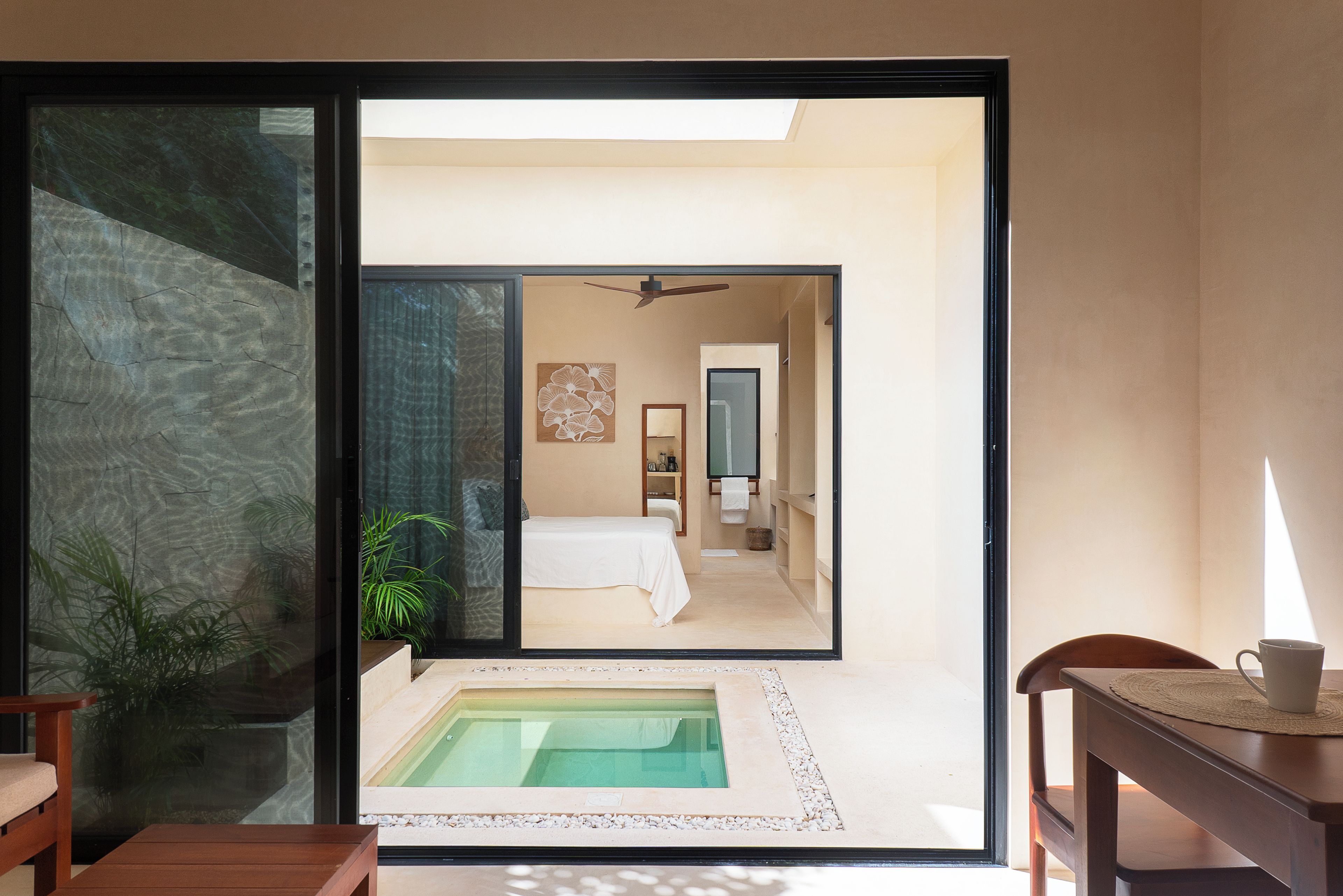 Una habitación de hotel moderna con puerta de vidrio que da a un espacio exterior privado, una cama, un baño pequeño y una zona de estar con una mesa y una silla.