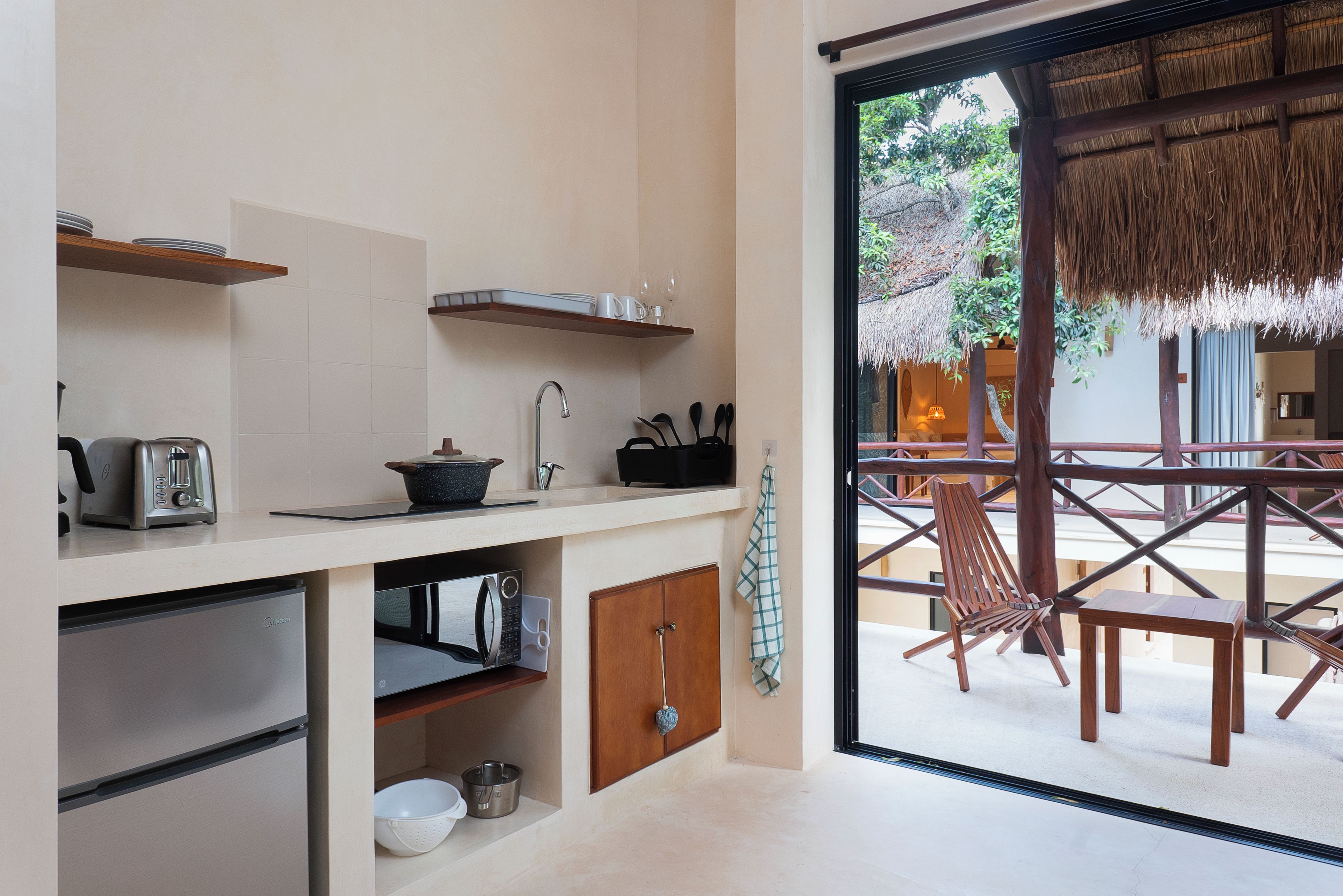 A modern kitchen with a built-in oven, microwave, and sink. There is a dining area with a thatched roof and wooden furniture visible through the glass door.