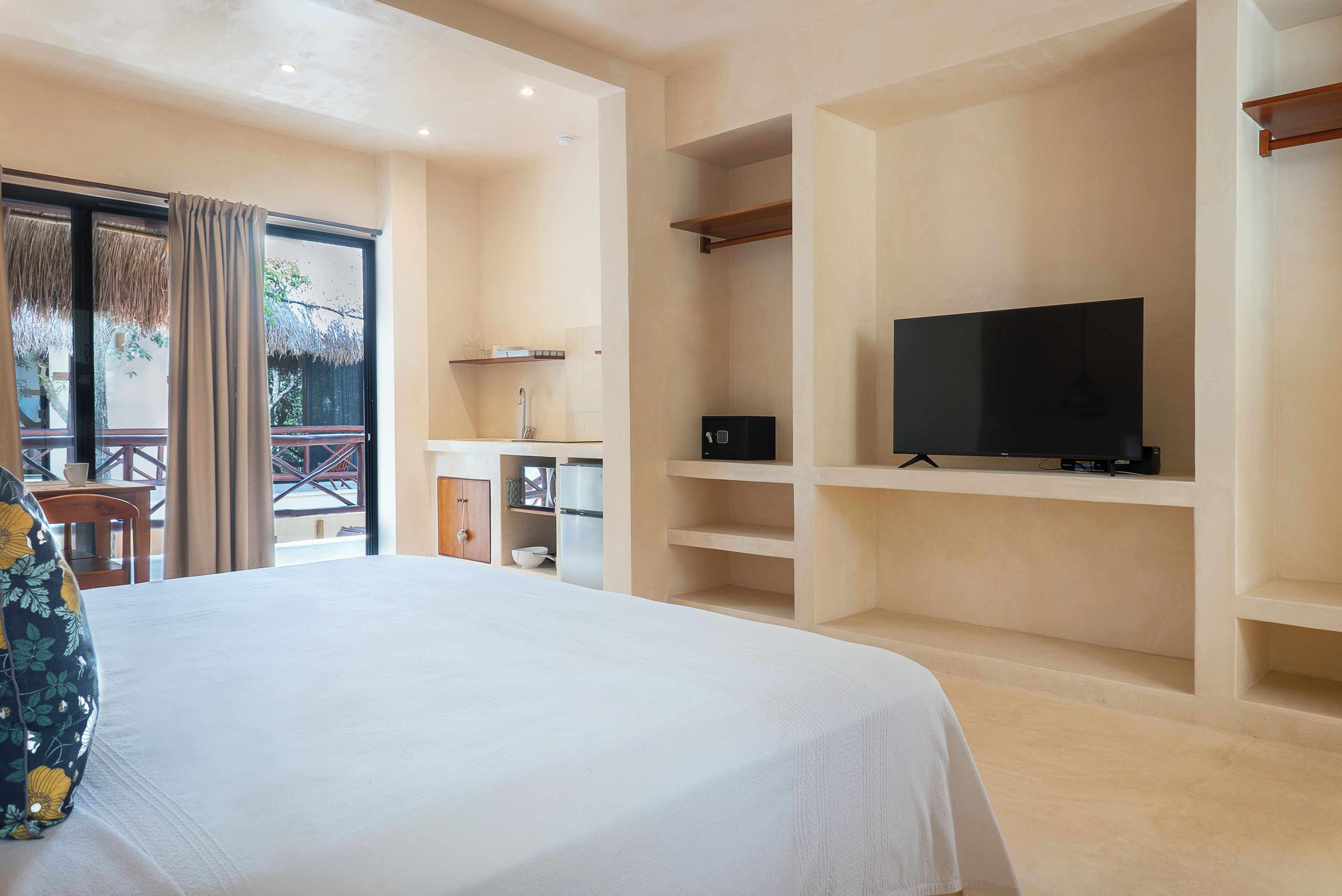 A hotel room with a bed, a television on a shelf, a small kitchen area, and a balcony with a table and chairs.